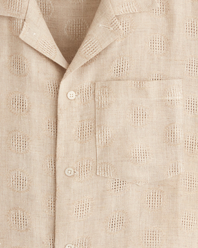 The Acorn Cap Shirt, a beige, short-sleeve button-up with a textured dot pattern, hangs on a metal hanger against a white tiled wall, showcasing its one-of-a-kind appeal.