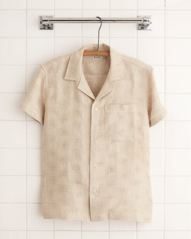 The Acorn Cap Shirt, a beige, short-sleeve button-up with a textured dot pattern, hangs on a metal hanger against a white tiled wall, showcasing its one-of-a-kind appeal.