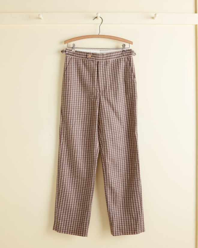 The Acorn Check Trousers - 28 hang gracefully on a wooden hanger against a cream wall, showcasing their timeless charm.