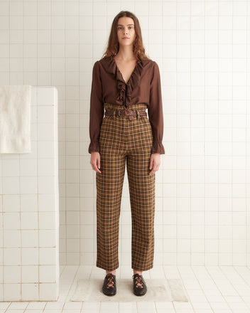 A person stands in a tiled room wearing a brown ruffled blouse and Ardara Check Shakey Trousers, paired with black shoes.