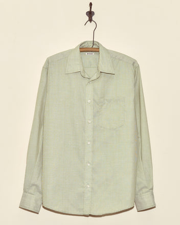 The Algae Ripple Shirt - M, a light green long-sleeved button-up made from deadstock shirting fabric, hangs on a hook against a beige wall.