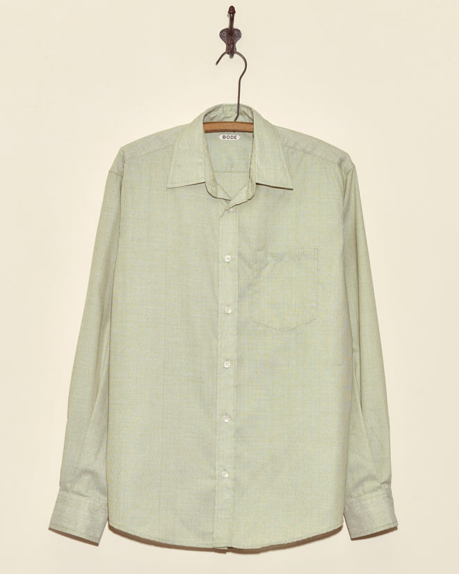 The Algae Ripple Shirt - M, a light green long-sleeved button-up made from deadstock shirting fabric, hangs on a hook against a beige wall.