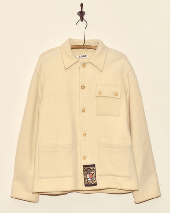 The Almond Cream Jacket – S/M, a unique cream-colored button-up with pockets made in New York, hangs on a hook against a plain wall.