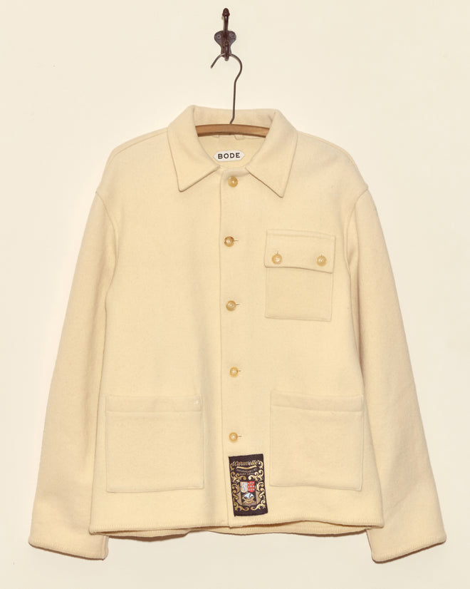 The Almond Cream Jacket – S/M, a unique cream-colored button-up with pockets made in New York, hangs on a hook against a plain wall.