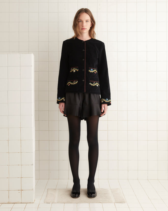 A person in a black outfit stands in a tiled room on a small white rug, wearing the elegant Alpineflower Rice Jacket adorned with a wildflower-and-vine pattern.