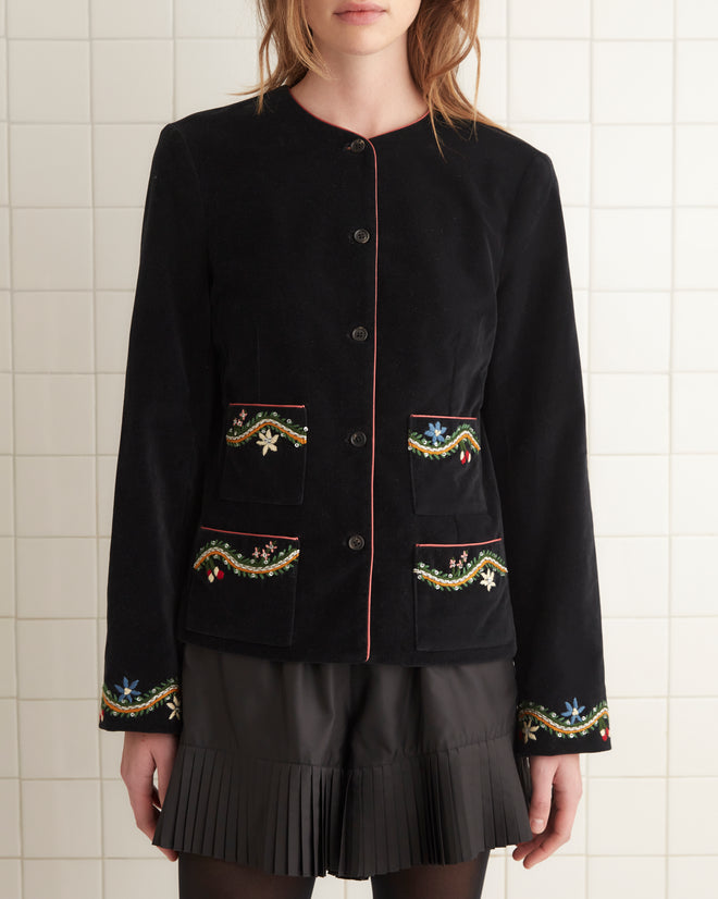 A person in a black outfit stands in a tiled room on a small white rug, wearing the elegant Alpineflower Rice Jacket adorned with a wildflower-and-vine pattern.