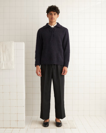 A person wearing the Alpine Pullover in navy, featuring a lace-up neckline, stands in a tiled room; black pants complete the look, while a towel hangs on the wall.