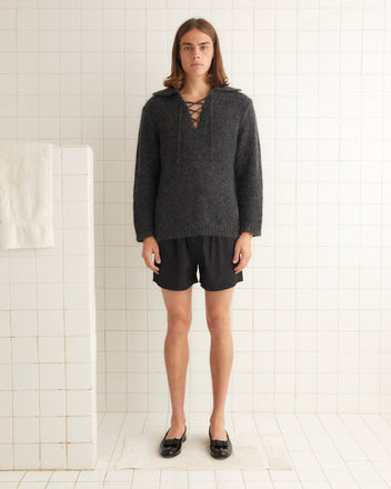 A person is standing in a bathroom wearing the Alpine Pullover - Grey, paired with black shorts and black shoes, with a white towel hanging on the tiled wall.