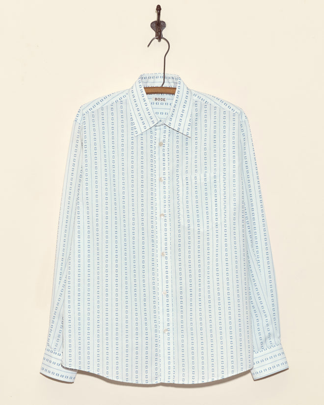 The Alpine Row Shirt - M is a light blue long-sleeve button-up, made from vintage shirting fabric with vertical blue and white dotted stripes, and hangs on a hook.