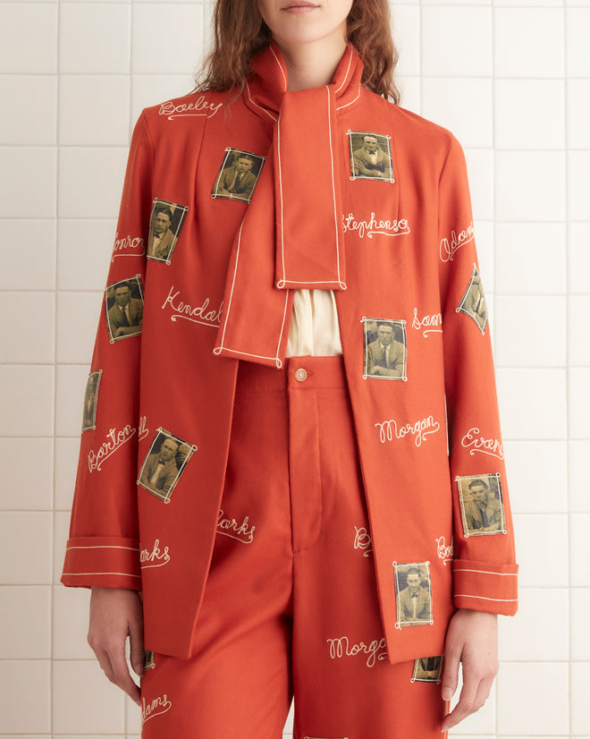 A person wearing an elongated fit, scarlet wool suit and black shoes stands in a tiled room on a white mat, featuring the Alumni Jacket.