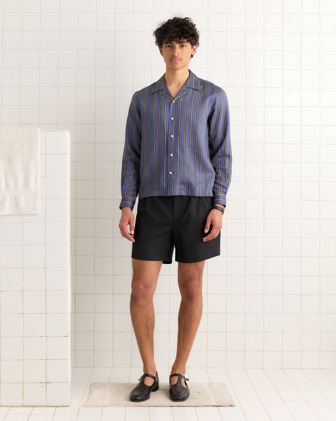 A man stands in a tiled room wearing the Alumni Stripe Long Sleeve Shirt - Blue, inspired by 1960s-1980s style, paired with black shorts and black shoes.