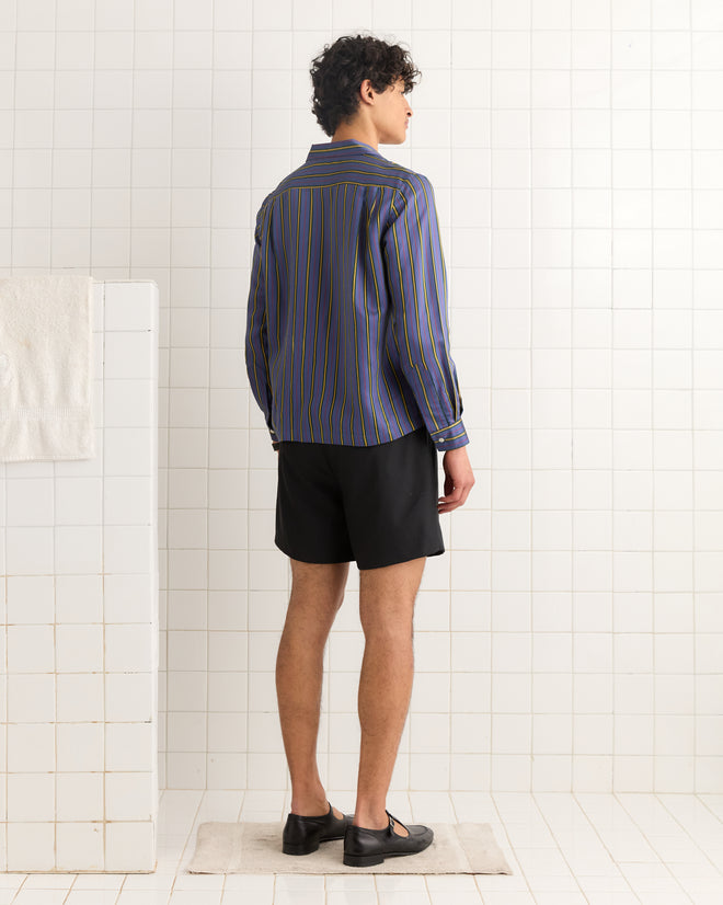 A man stands on a mat in a tiled room, wearing the Alumni Stripe Long Sleeve Shirt in Blue, inspired by 1960s-1980s style and crafted from a viscose silk mix, paired with black shorts and black shoes.