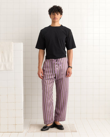 A person stands in a tiled room wearing a black t-shirt, Alumni Stripe Pajama Pants- Purple, and black shoes.