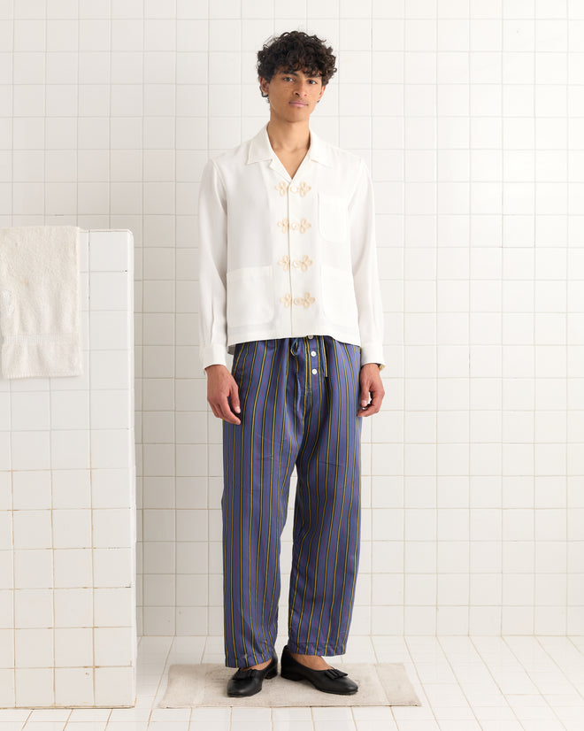 A person stands in a tiled room wearing a white shirt and Alumni Stripe Pajama Pants- Blue with a bold stripe pattern, paired with black shoes.