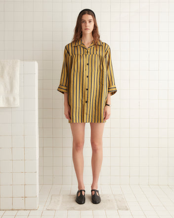 A woman with long hair stands in a tiled room, wearing the Alumni Stripe Quincy Dress, which features vintage-inspired yellow and black stripes. The silky fabric catches the light beautifully as she stands barefoot on a mat, her hands resting calmly by her sides.