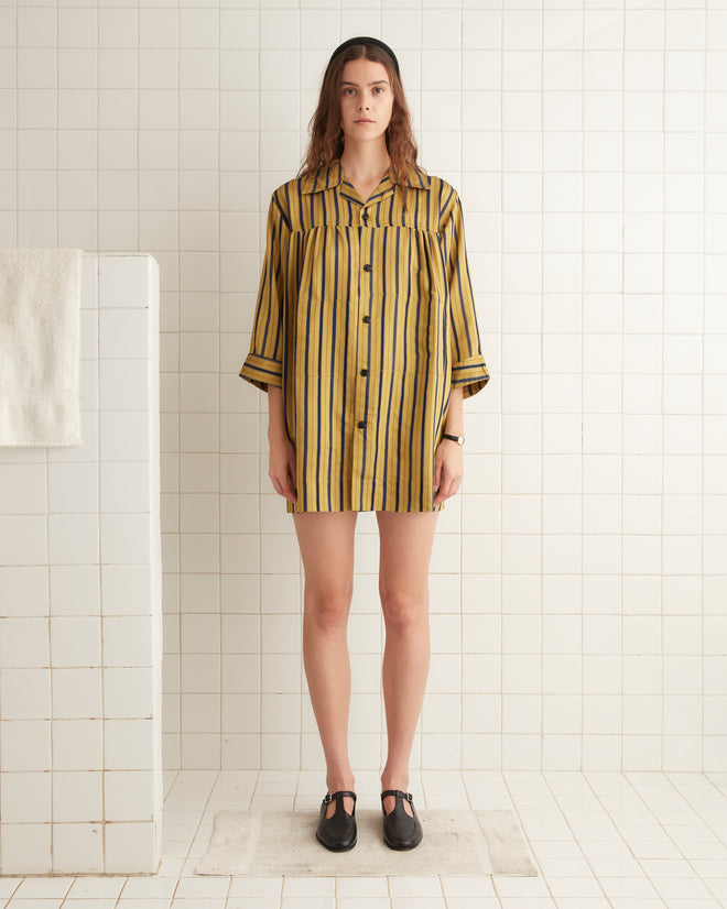 A woman with long hair stands in a tiled room, wearing the Alumni Stripe Quincy Dress, which features vintage-inspired yellow and black stripes. The silky fabric catches the light beautifully as she stands barefoot on a mat, her hands resting calmly by her sides.