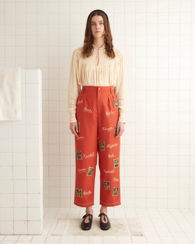 A woman wearing a cream blouse and midcentury-inspired Alumni Trousers in scarlet wool, standing in a tiled room adorned with text and images.