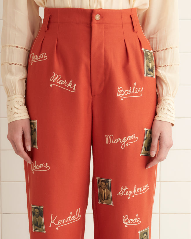 A woman wearing a cream blouse and midcentury-inspired Alumni Trousers in scarlet wool, standing in a tiled room adorned with text and images.