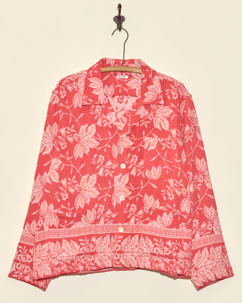 The Amaryllis Long Sleeve Shirt - L, featuring a unique red design with a white floral pattern inspired by damask, hangs on a hook against a plain wall.