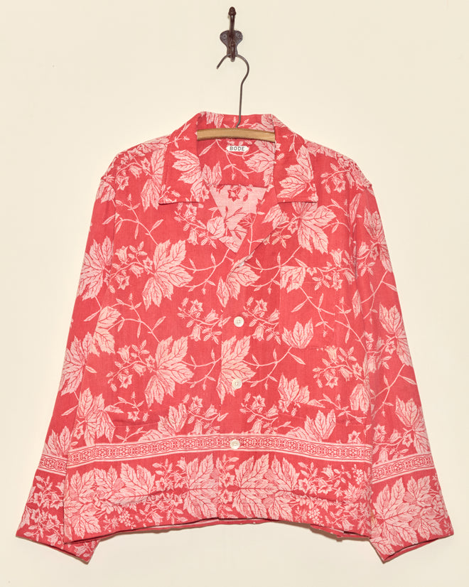 The Amaryllis Long Sleeve Shirt - L, featuring a unique red design with a white floral pattern inspired by damask, hangs on a hook against a plain wall.