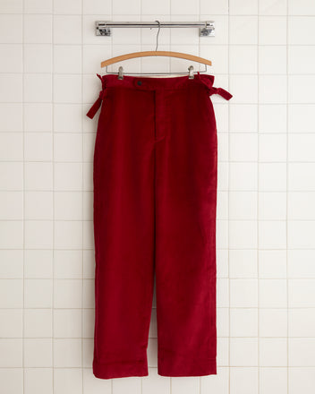 The Amor Corduroy Trousers, featuring a chic tie belt, hang elegantly on a wooden hanger against a tiled wall, showcasing the perfect blend of comfort and style.