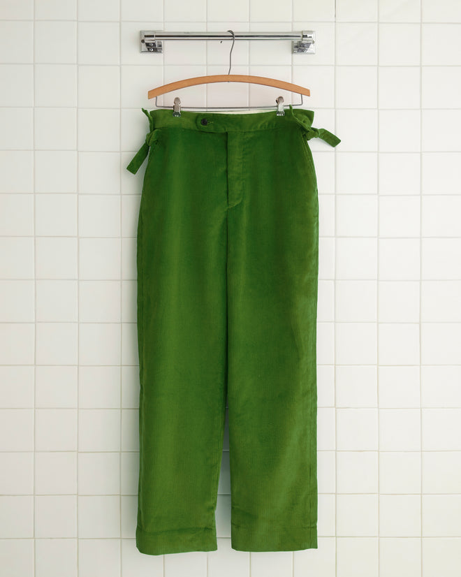 The Shamrock Green Trousers, crafted in New York, hang on a wooden hanger against a white tiled wall, showcasing their unique side-tie adjusters and embodying both style and craftsmanship.