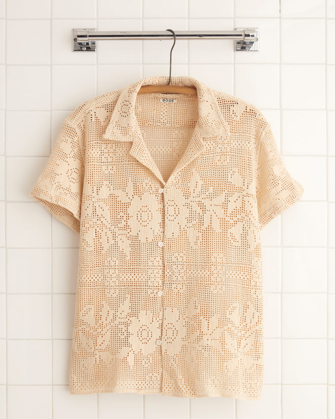 The Anemone Panel Shirt - S, a beige, short-sleeve vintage piece with a floral filet lace pattern, hangs on a metal hanger against a tiled wall, making it truly one-of-a-kind.