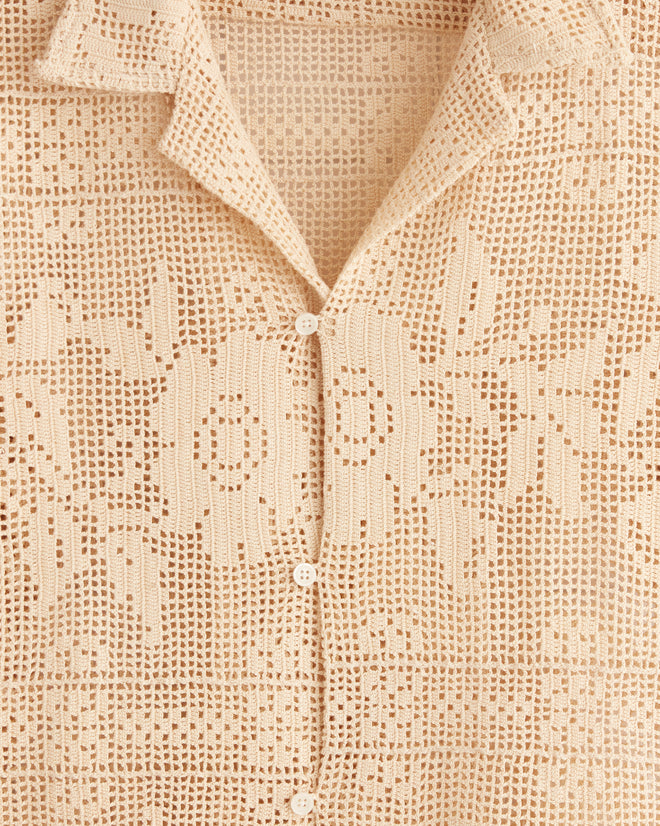 The Anemone Panel Shirt - S, a beige, short-sleeve vintage piece with a floral filet lace pattern, hangs on a metal hanger against a tiled wall, making it truly one-of-a-kind.