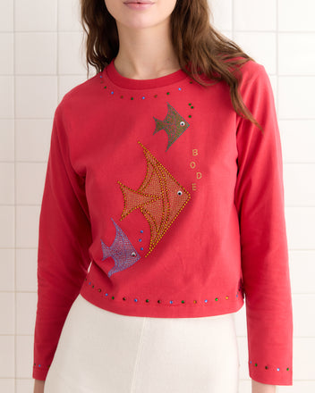 A woman in the Angel Fish Tee - Red, featuring an angel fish design with shimmering rhinestones on 100% cotton, stands gracefully against a tiled wall.