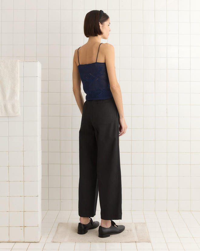 A person with short hair stands in a tiled bathroom, wearing the Angel Fish Tank - Navy and black pants, facing away.