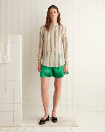 A woman stands in a tiled room wearing an Antique Stripe Shirt styled in a 1920s fashion, green pleated shorts, and black shoes.