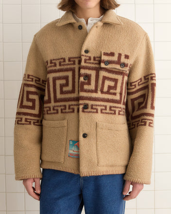 A person stands against a tiled wall wearing the Apollo Ornament Jacket - M/L, a unique wool piece with geometric brown patterns inspired by the Greek meander motif, paired with blue jeans.