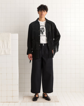 A person in black wide-leg pants, the Appalachian Jacket - Black with 1960s Western fringe details, and a printed white T-shirt stands against a tiled wall.