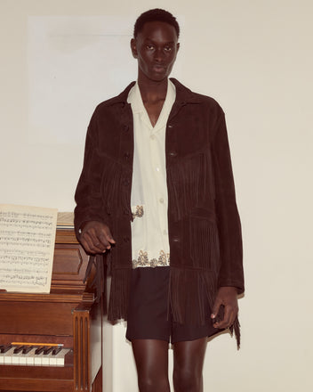 Wearing the Appalachian Jacket - Brown, a person in Western attire with hand-cut fringe stands by a piano with sheet music, facing the camera.