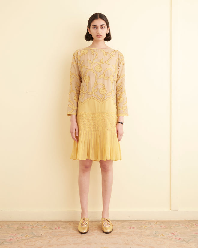 A person stands indoors against a light-colored wall, wearing the Appliqué Harlayne Dress, which features long sleeves, a smocked drop waist, intricate yellow designs, and gold shoes.