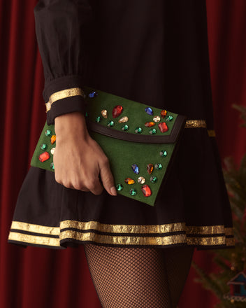 The person carries the Gem Venue Clutch in Green/Black, a suede envelope adorned with colorful gems, while wearing a black dress featuring gold trim and fishnet stockings.