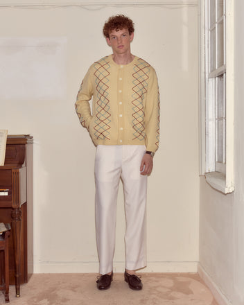 A man in a yellow Beaded Argyle Sketch Cardigan and white pants stands indoors near a window and a piano.