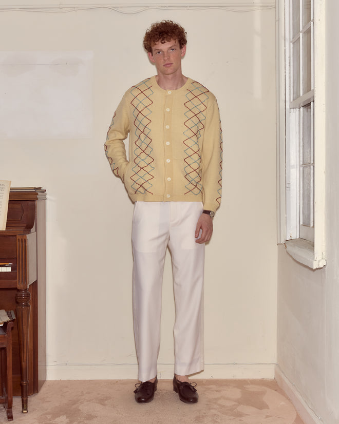 A man in a Beaded Argyle Sketch Cardigan with antique pearl buttons and white pants stands indoors near a piano and window.