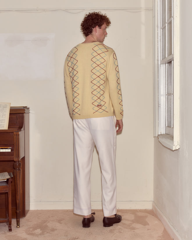 Man with curly hair wearing the Beaded Argyle Sketch Cardigan with antique pearl buttons and white pants stands by a wall near a window and piano.