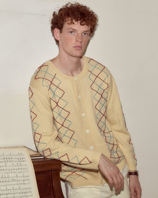 A young man in the Beaded Argyle Sketch Cardigan with pearl buttons leans on a piano with sheet music, looking at the camera.