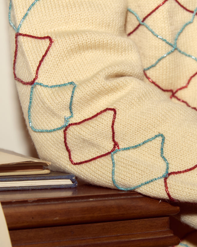The Beaded Argyle Sketch Cardigan, featuring red and blue stitched square patterns on cream knitwear, is draped over books on a wooden surface.