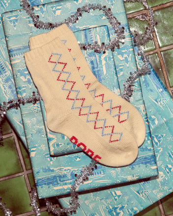 Argyle Sketch Socks in cream merino wool feature a red and blue diamond pattern, shown on blue gift wrap with silver tinsel.