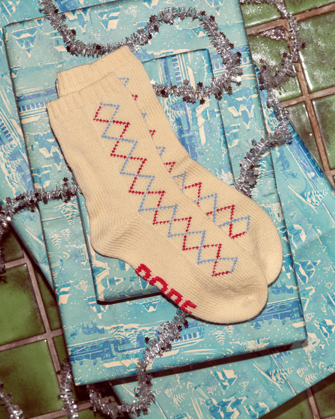 Argyle Sketch Socks in cream merino wool feature a red and blue diamond pattern, shown on blue gift wrap with silver tinsel.