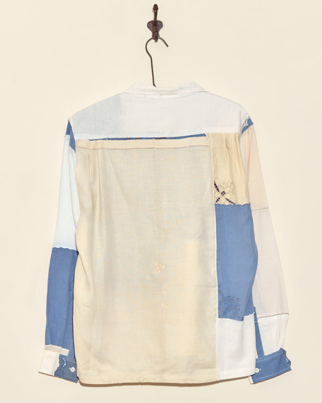 The Aster Napkin Shirt - M/L, a unique long-sleeve vintage piece in blue, cream, and white, hangs on a hook against a plain wall.