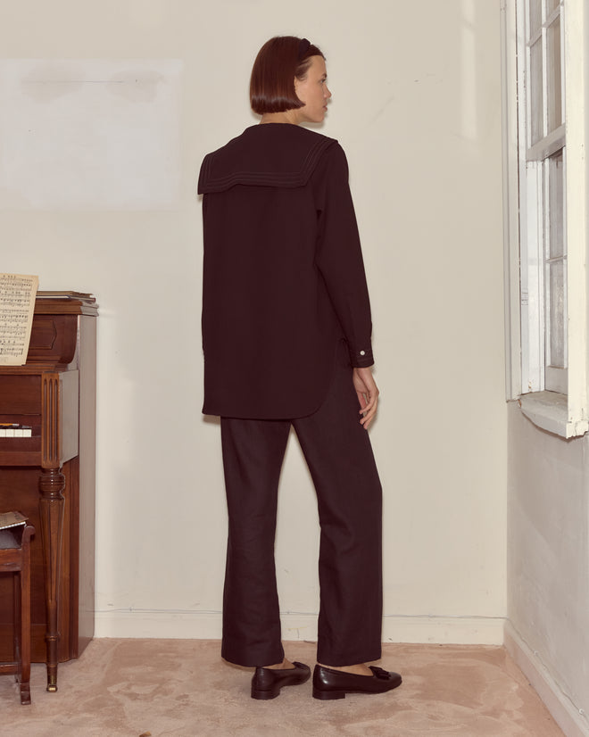 A woman wearing the Astor Blouse stands by a piano, facing away from the camera near a bright window.