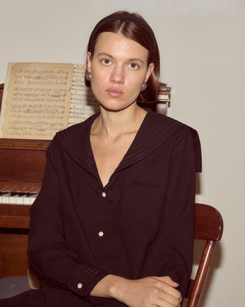 A woman in the Astor Blouse sits by a piano with sheet music, looking at the camera with a neutral expression.