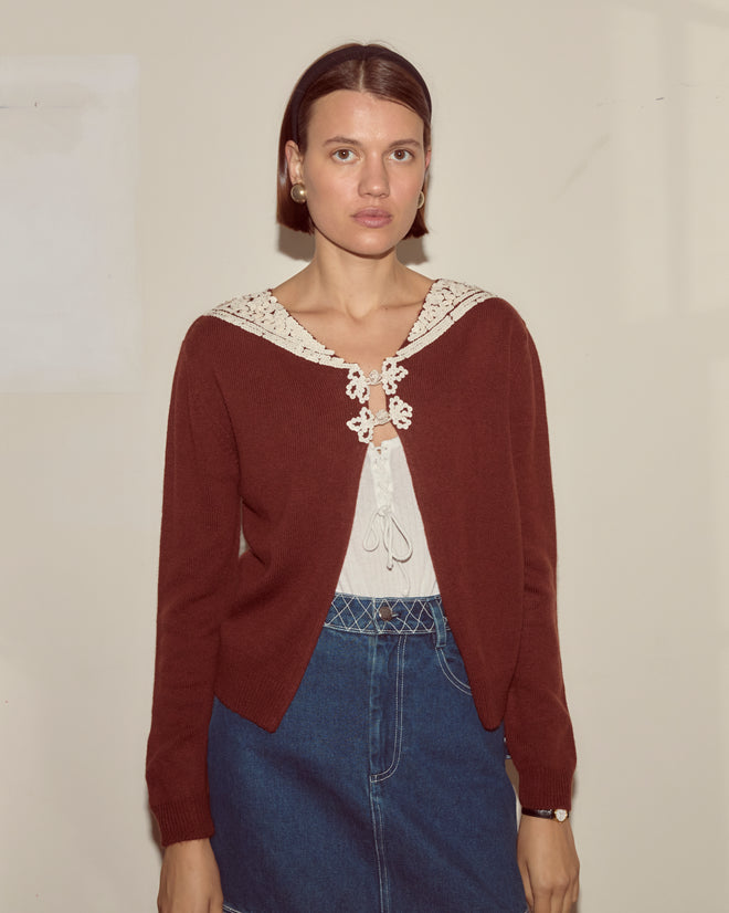 Woman wears the Astoria Cardigan in brown over a white blouse and denim skirt, standing against a beige wall.