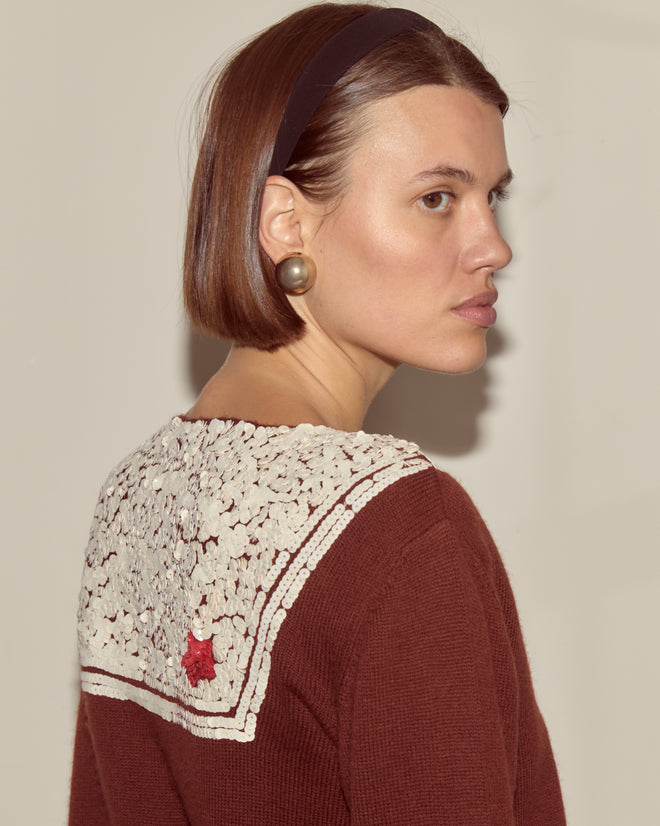Woman with a bob haircut, pearl earring, headband, and the Astoria Cardigan in brown featuring a white lace collar and 1970s-inspired knitwear style with a red star.