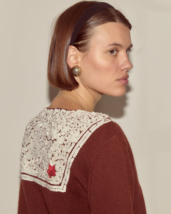 Woman with a bob haircut, pearl earring, headband, and the Astoria Cardigan in brown featuring a white lace collar and 1970s-inspired knitwear style with a red star.
