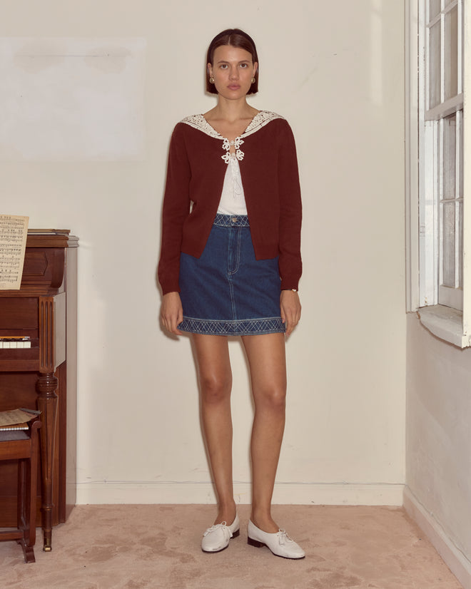 A woman wearing the Astoria Cardigan in brown, paired with a white top and denim skirt, stands indoors beside a piano and window.